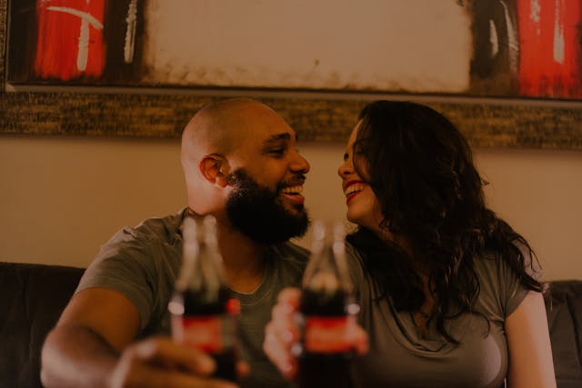 Stel zit dicht bij elkaar met een fles cola en lacht met gezichten dicht bij elkaar.