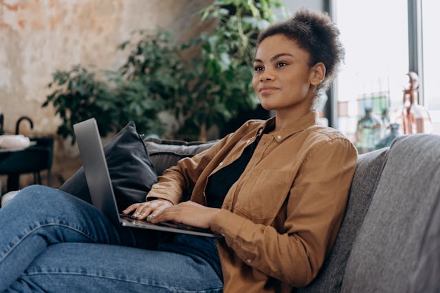 Vrouw zit met laptop op schoot en kijkt nadenkend in de verte.