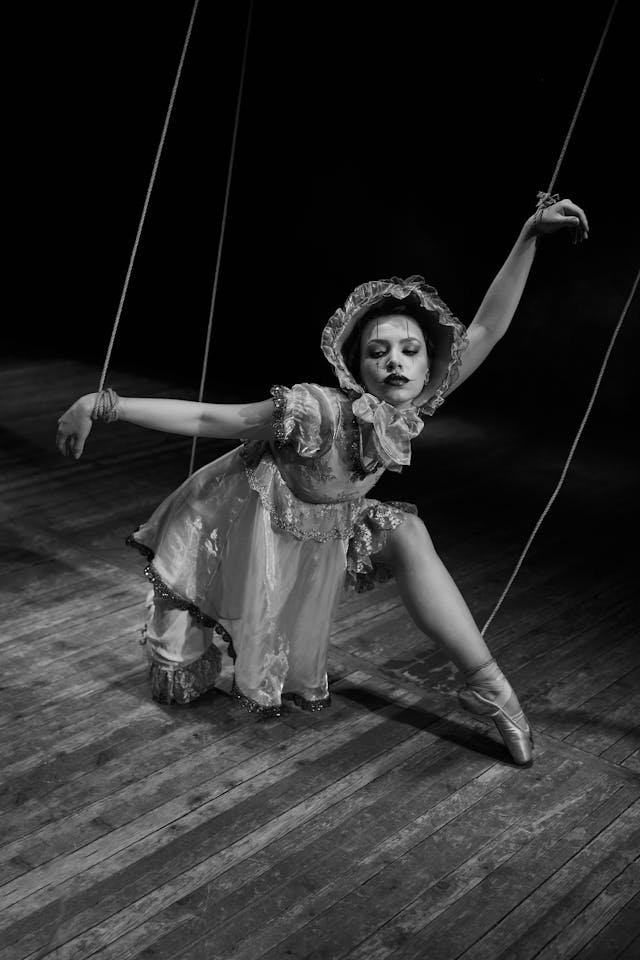 Zwart-wit foto van een balletdanseres verkleed als marionet met touwtjes aan polsen en enkels.