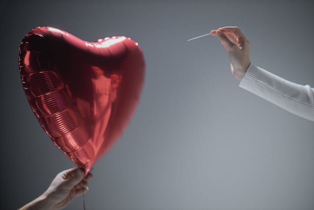 Hand met een speld naast een hartvormige ballon, symbool voor de moeilijke maar noodzakelijke keuze na een relatiebreuk.