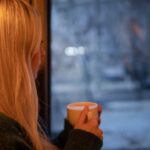 Vrouw die peinzend met een kop koffie uit het raam kijkt naar een besneeuwd landschap.