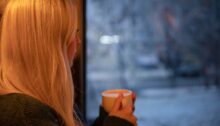 Vrouw die peinzend met een kop koffie uit het raam kijkt naar een besneeuwd landschap.