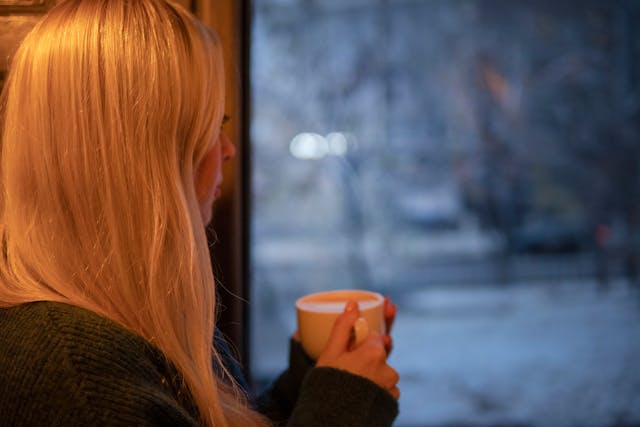 Vrouw die peinzend met een kop koffie uit het raam kijkt naar een besneeuwd landschap.
