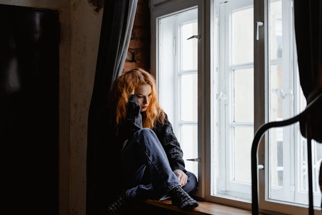 Vrouw zit alleen in de vensterbank met een hand in haar haar en kijkt neerslachtig naar beneden, symbool voor eenzaamheid na een relatiebreuk.